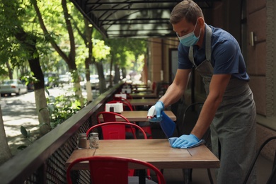 Photo of Waiter in mask and gloves disinfecting table at outdoor cafe