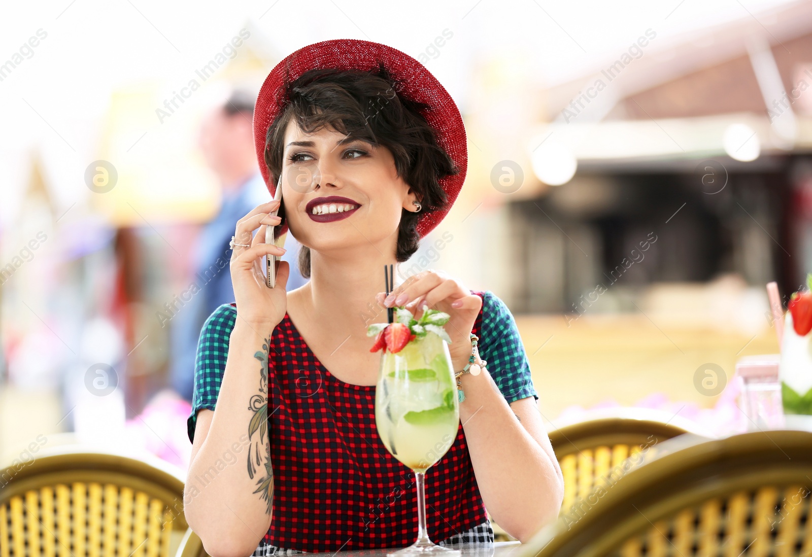 Photo of Young woman with glass of tasty lemonade talking on phone in open-air cafe