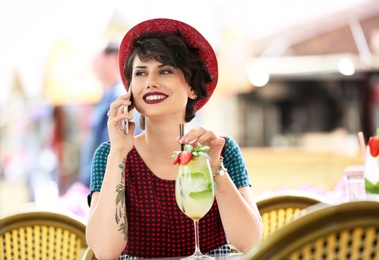 Young woman with glass of tasty lemonade talking on phone in open-air cafe