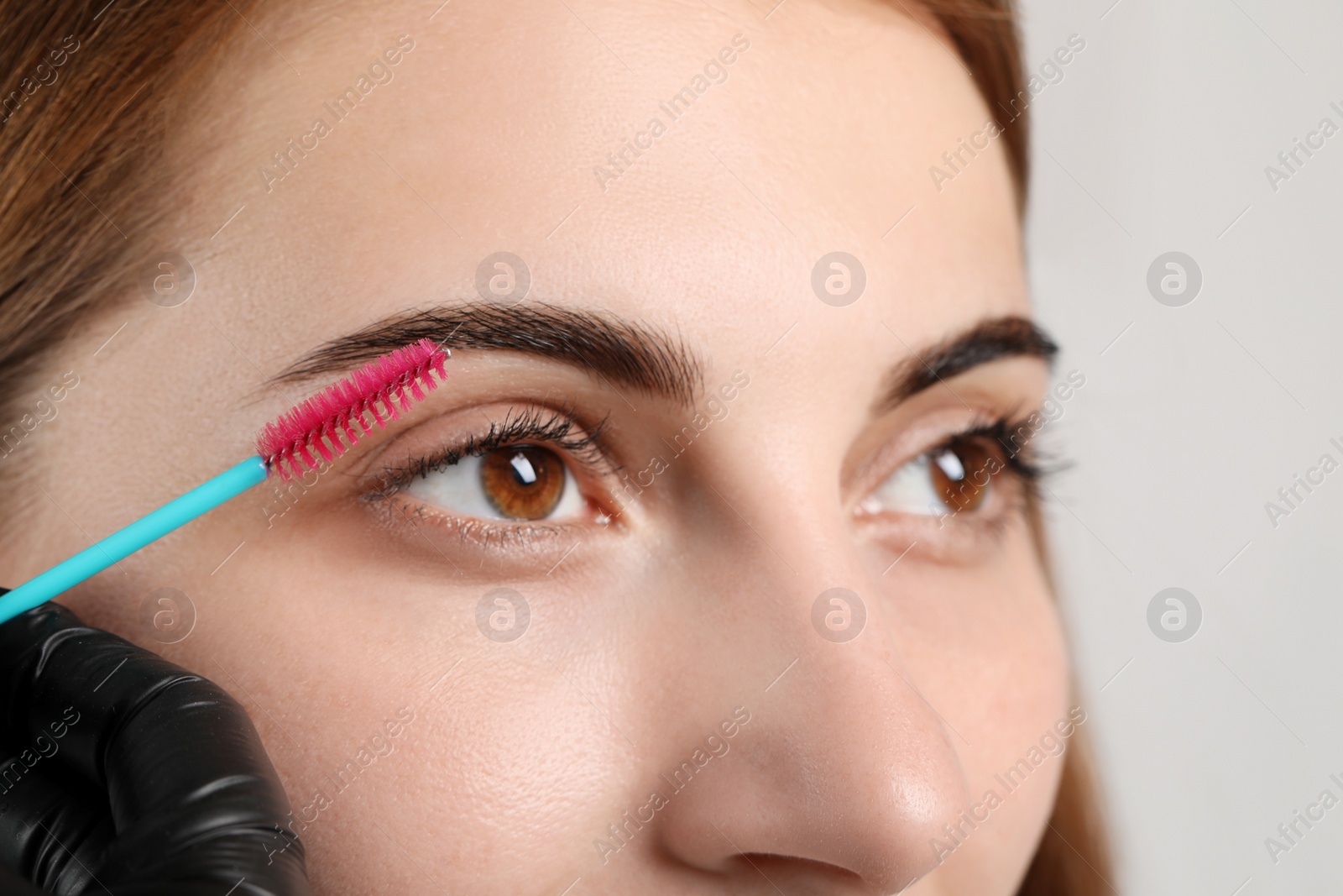 Photo of Beautician brushing woman's eyebrows after tinting on grey background, closeup