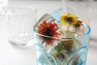 Glass of ice cubes with flowers on blurred background, closeup