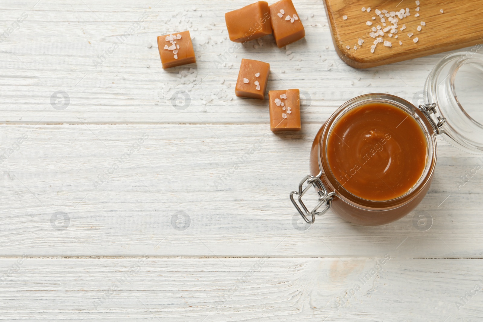 Photo of Yummy salted caramel in glass jar and candies on white wooden table, flat lay. Space for text