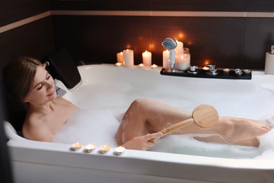 Beautiful woman rubbing leg with brush while taking bubble bath indoors. Romantic atmosphere
