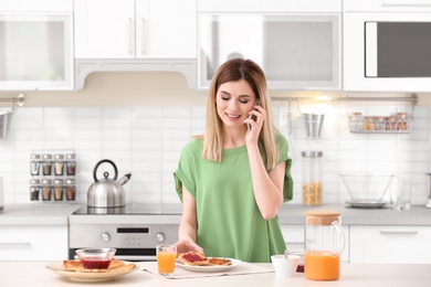 Photo of Beautiful woman eating tasty toasted bread with jam while talking on mobile phone at table in kitchen