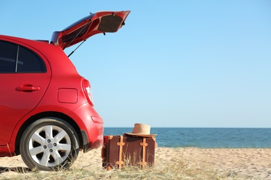 Photo of Modern car, retro suitcase and hat on sand near sea, space for text. Beach accessories