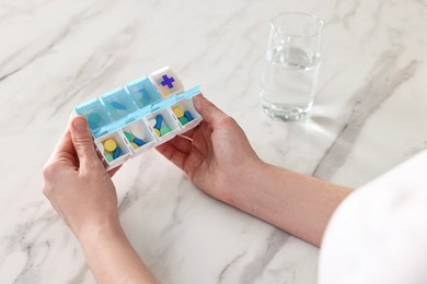 Photo of Woman with pills, organizer and glass of water at white marble table, closeup