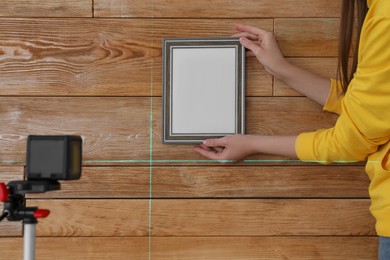 Photo of Woman using cross line laser level for hanging blank photo frame on wooden wall, closeup