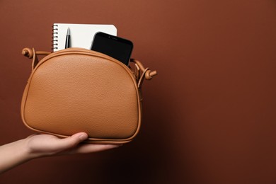 Photo of Woman holding stylish bag with smartphone and accessories on brown background, closeup. Space for text