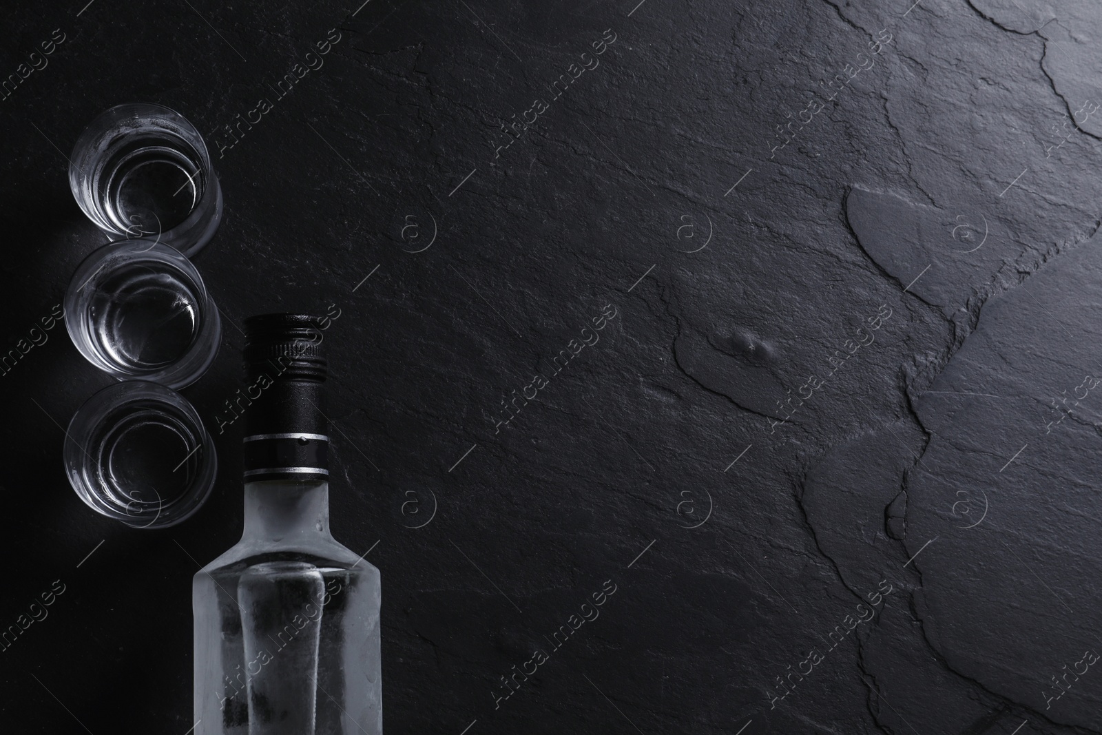 Photo of Bottle of vodka and shot glasses on black table, flat lay. Space for text