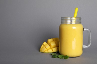 Photo of Mason jar of tasty smoothie with straw, mango and mint leaves on grey background. Space for text