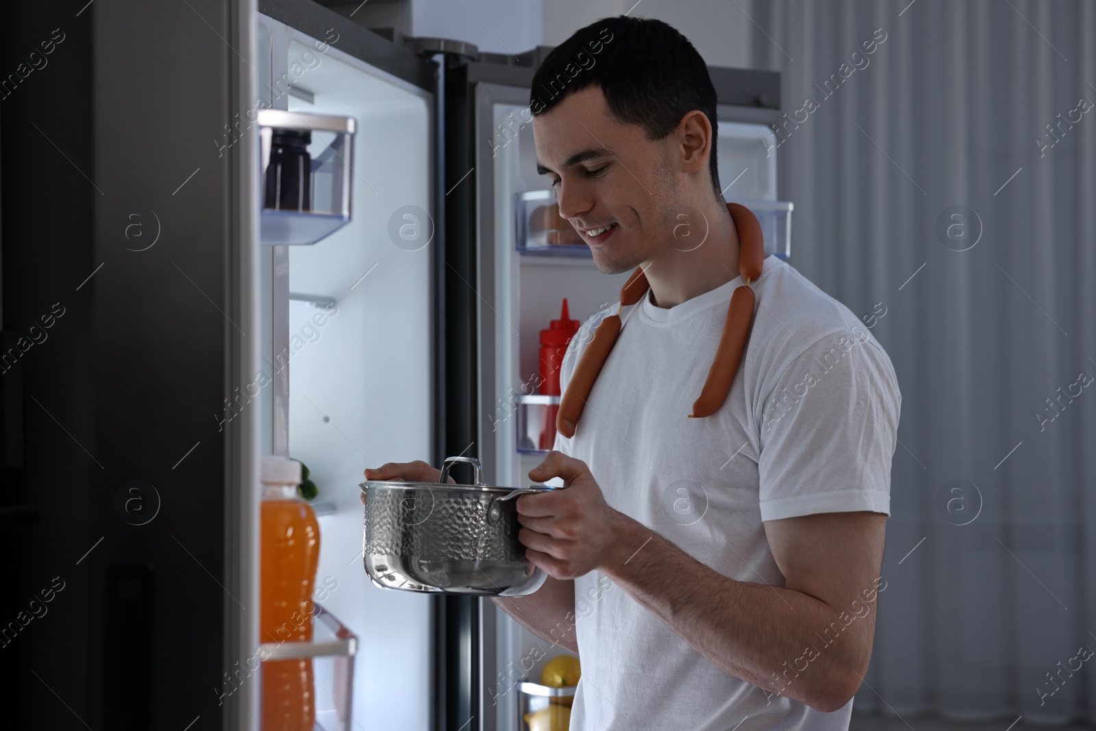 Photo of Man with sausages and pot near refrigerator in kitchen at night