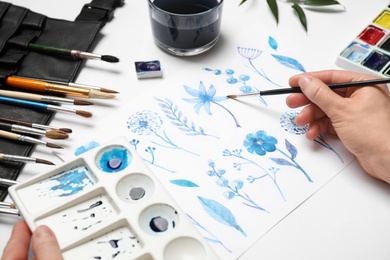 Photo of Woman painting flowers with watercolor at white table, closeup