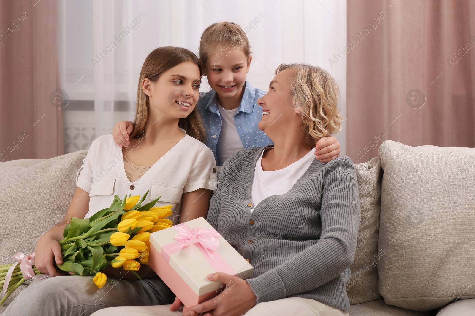 Photo of Little girl congratulating her mom and granny with flowers and gift at home. Happy Mother's Day