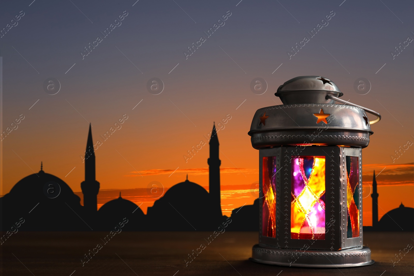 Image of Decorative Arabic lantern on wooden surface and silhouette of mosque at sunset on background, space for text