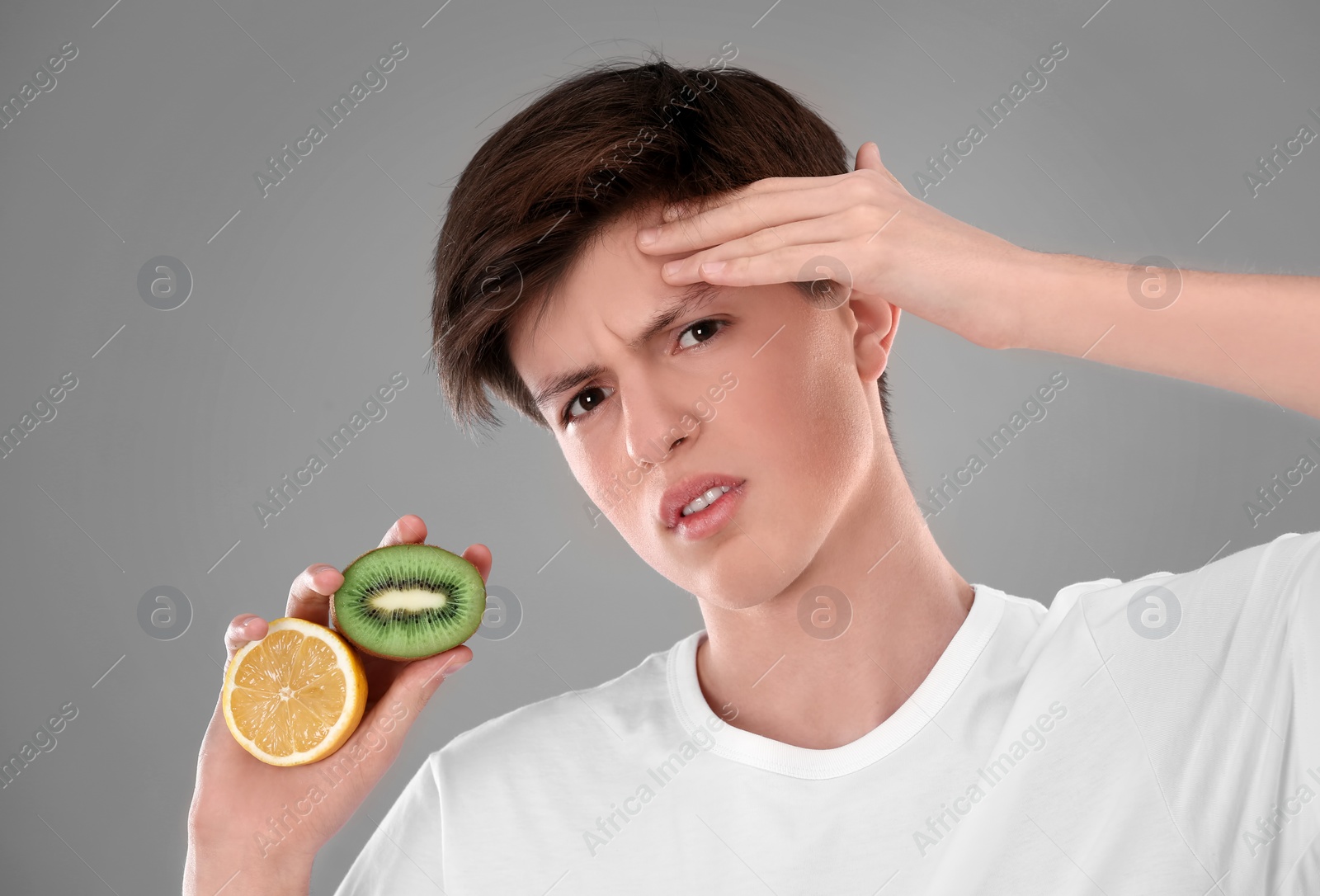 Photo of Teenage boy with acne problem holding lemon and kiwi on grey background. Skin allergy