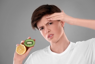 Teenage boy with acne problem holding lemon and kiwi on grey background. Skin allergy