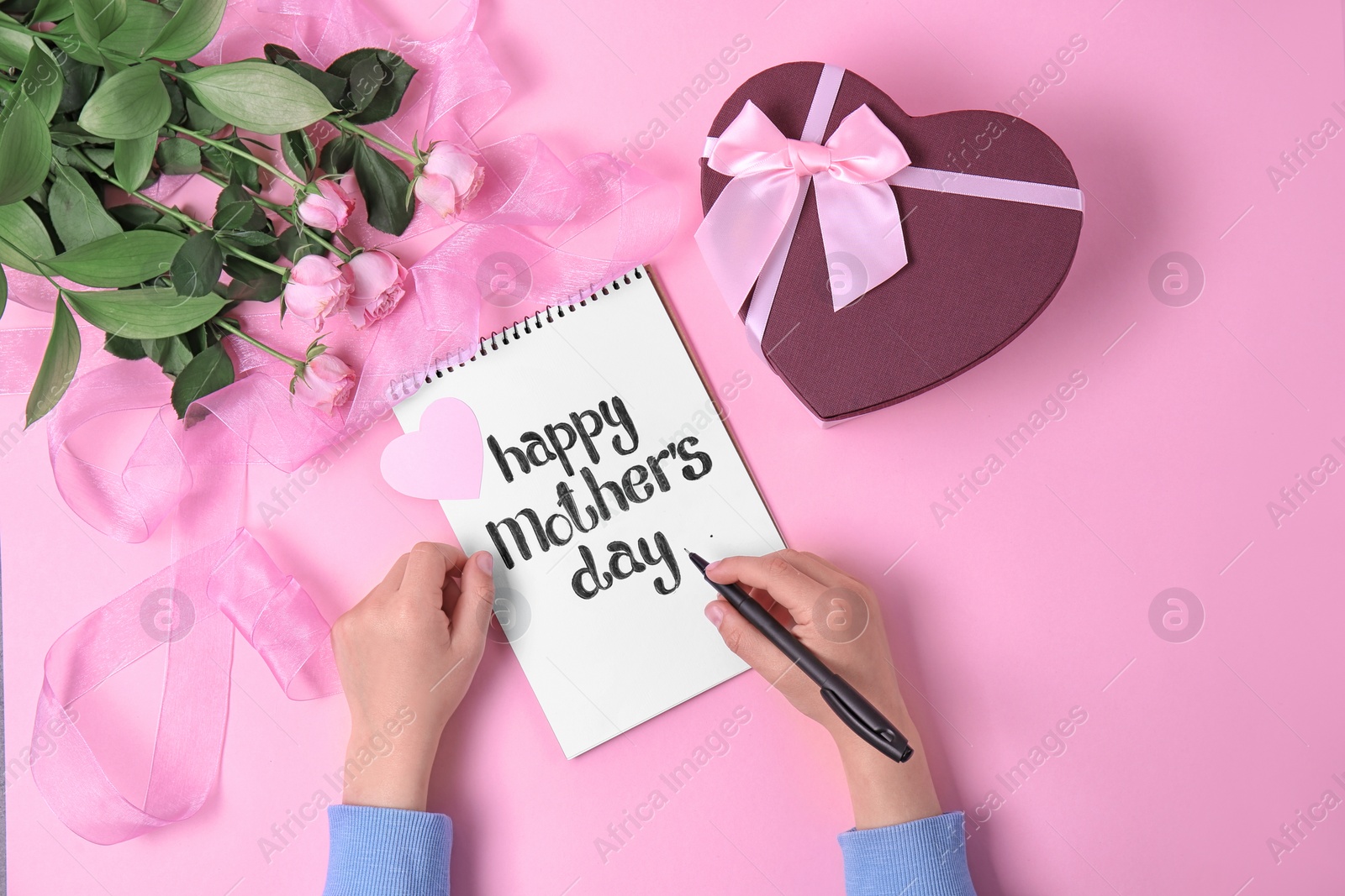 Photo of Child writing phrase "HAPPY MOTHER'S DAY" in notebook near gift and roses on table