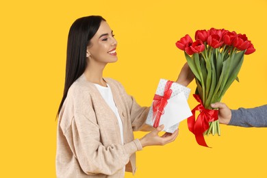 Happy woman receiving red tulip bouquet, greeting card and gift box from man on yellow background. 8th of March celebration