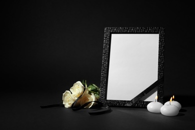 Photo of Funeral photo frame with ribbon, white roses and candles on black background. Space for design