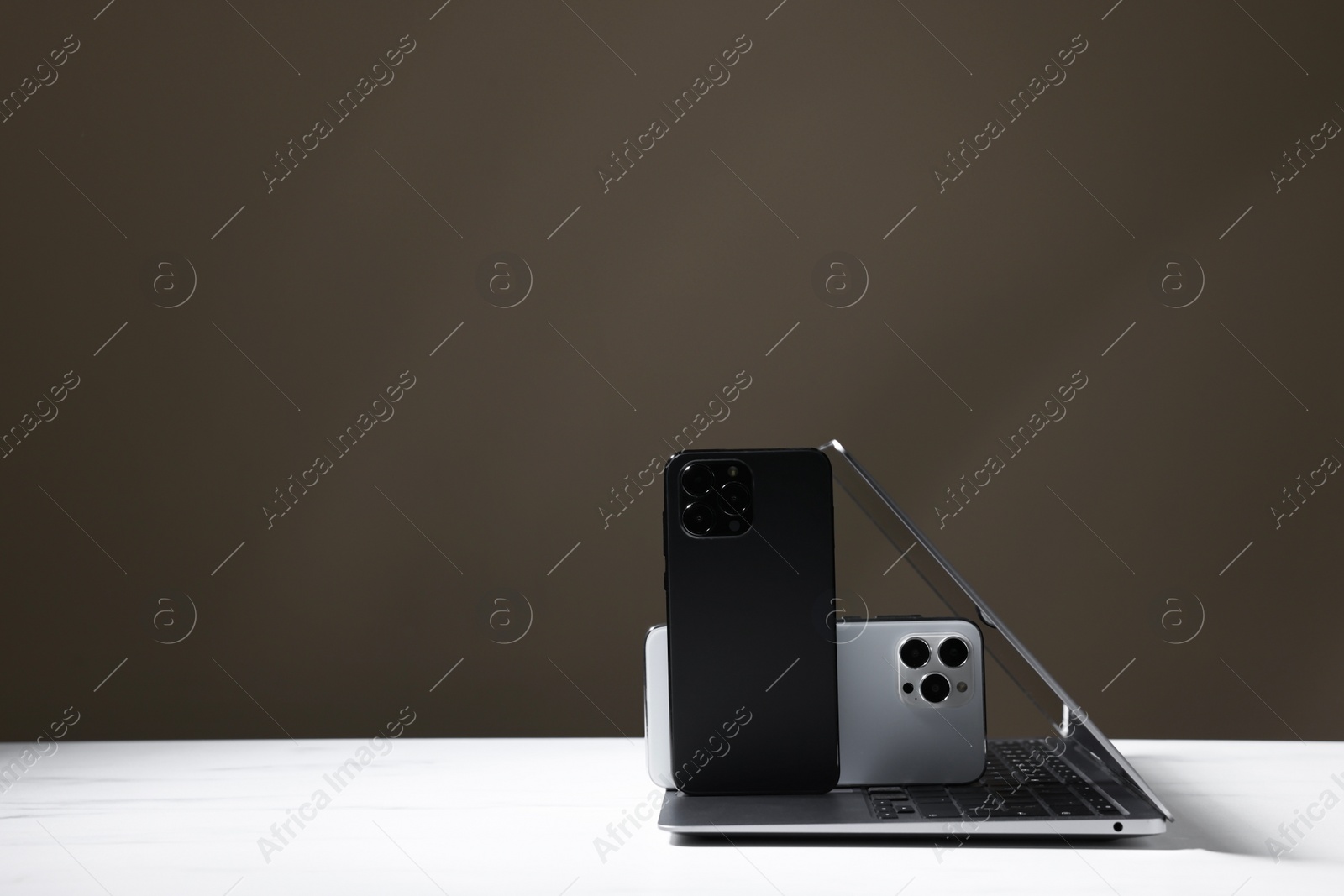 Photo of Modern laptop and smartphones on white table against brown background. Space for text