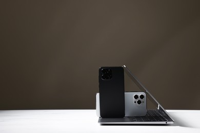 Photo of Modern laptop and smartphones on white table against brown background. Space for text