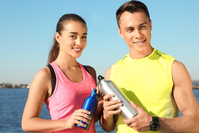 Young sporty couple with water bottles near river on sunny day