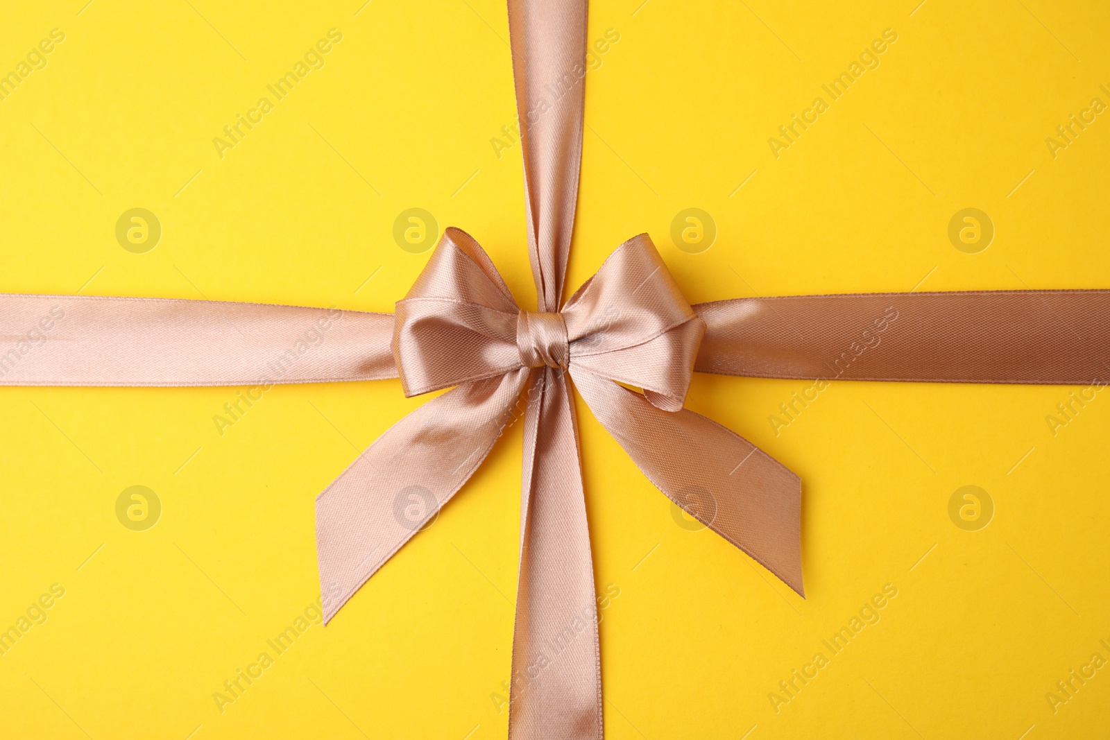 Photo of Beige satin ribbon with bow on yellow background, top view
