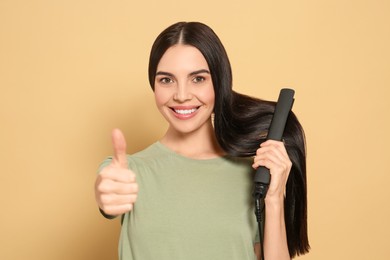 Beautiful happy woman showing thumbs up while using hair iron on beige background
