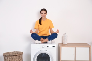 Young woman meditating on washing machine at home. Laundry day