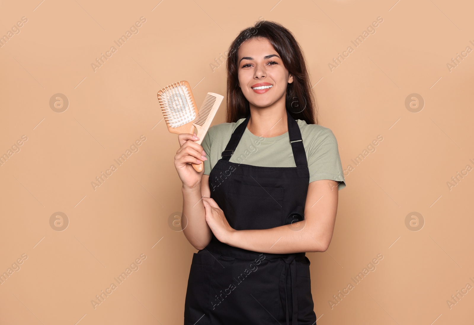 Photo of Happy professional hairdresser in apron with hairbrush and comb against pale orange background