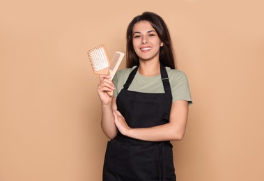 Photo of Happy professional hairdresser in apron with hairbrush and comb against pale orange background