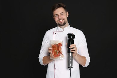 Smiling chef holding sous vide cooker and meat in vacuum pack on black background