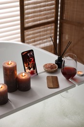 White wooden tray with smartphone, glass of wine and burning candles on bathtub in bathroom