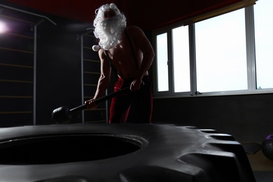 Photo of Young Santa Claus hitting heavy tire with hammer in modern gym