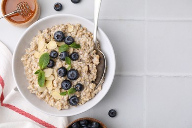 Photo of Tasty oatmeal with blueberries, mint and almond flakes in bowl on white tiled table, flat lay. Space for text