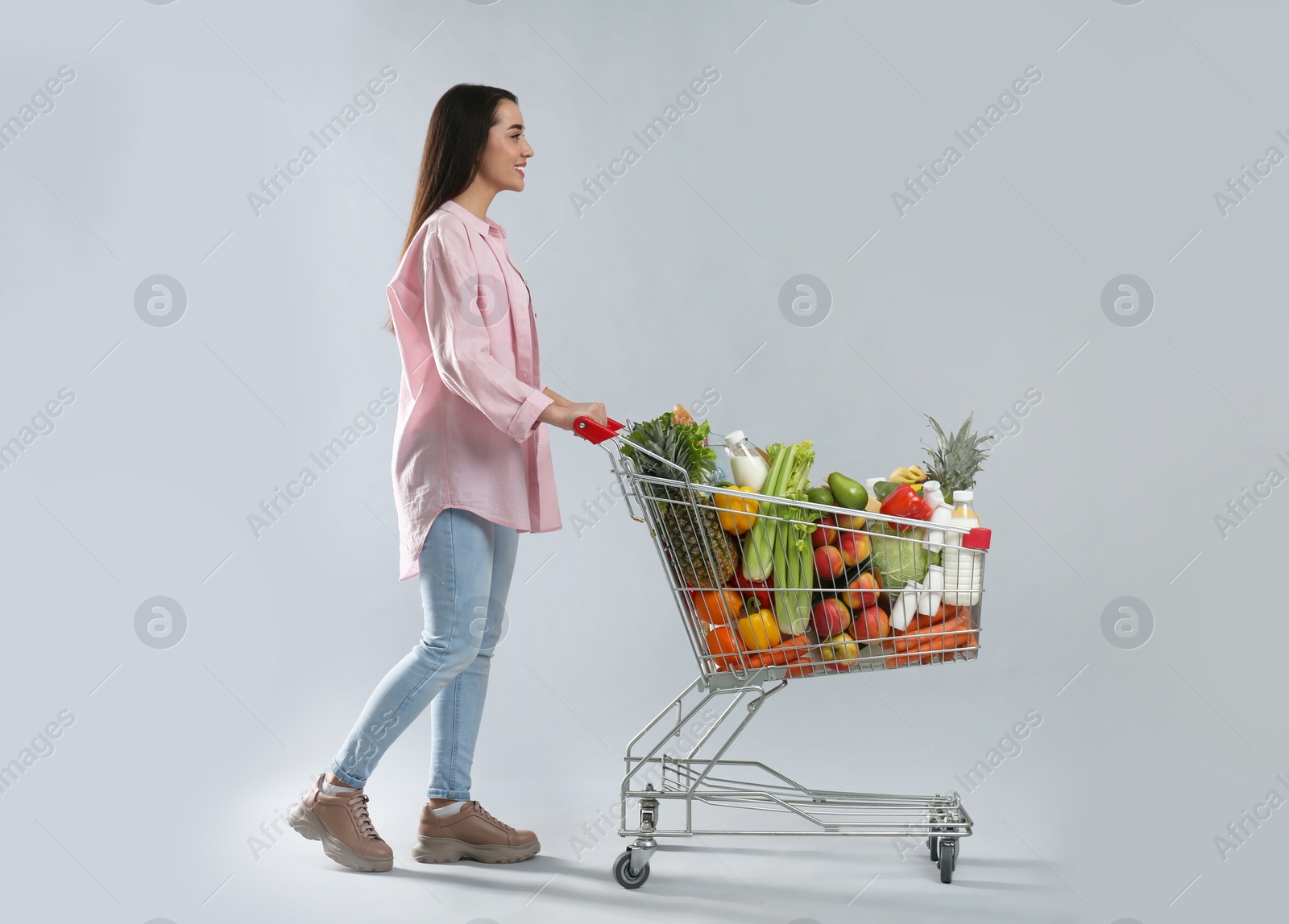 Photo of Young woman with shopping cart full of groceries on grey background