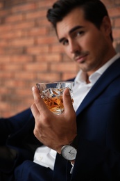 Young man with glass of whiskey near brick wall indoors