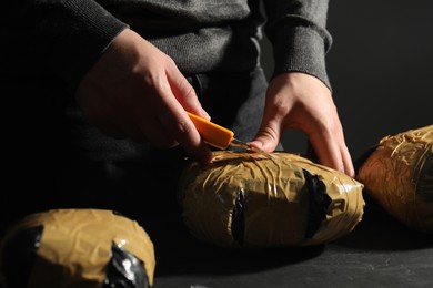 Photo of Smuggling and drug trafficking. Man opening package of narcotics with box cutter at dark table, closeup