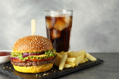 Photo of Burger with delicious patty, soda drink, french fries and sauce on dark table against gray background, closeup