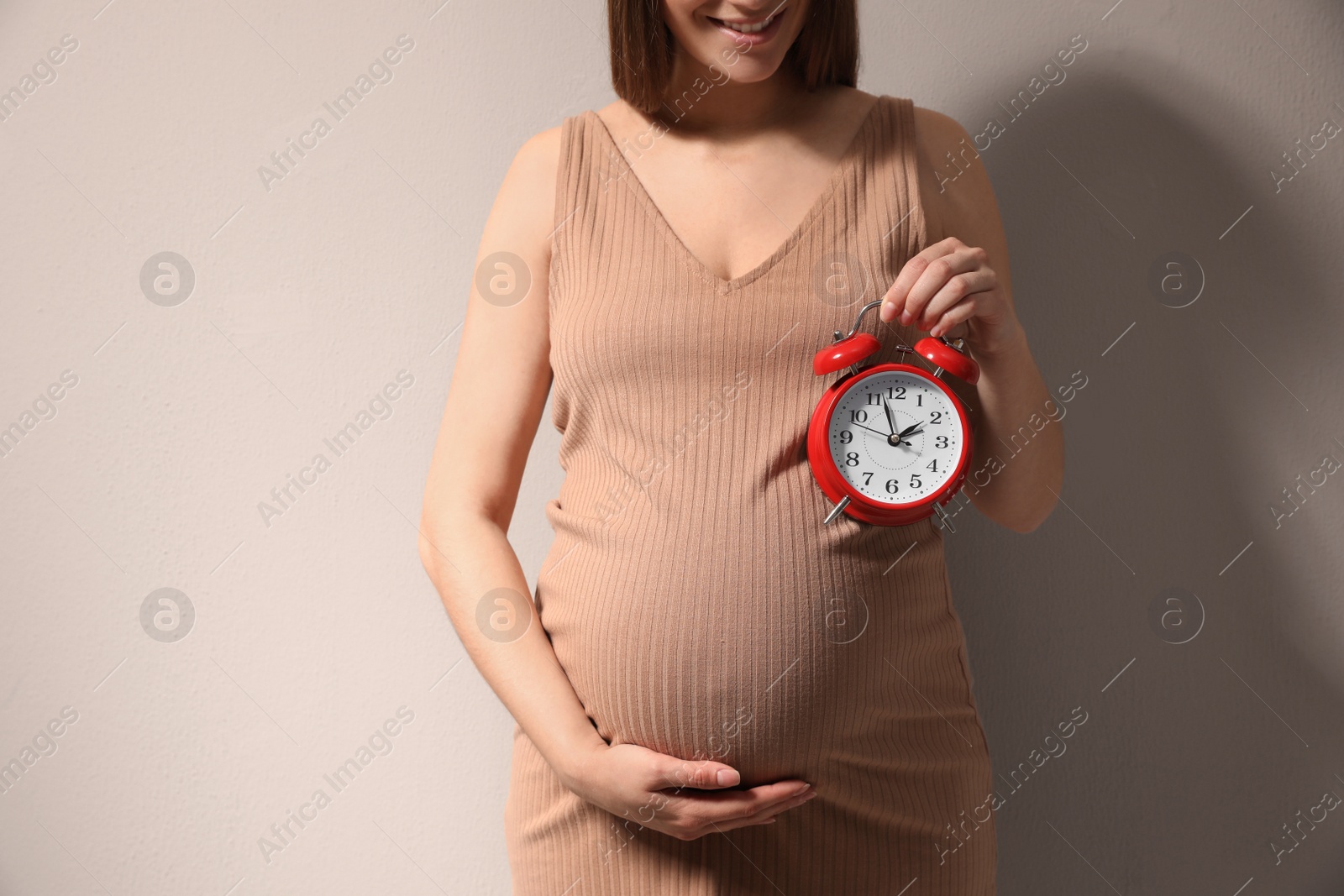 Photo of Young pregnant woman holding alarm clock near her belly on beige background, closeup. Time to give birth