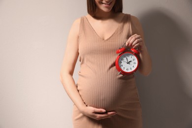 Young pregnant woman holding alarm clock near her belly on beige background, closeup. Time to give birth