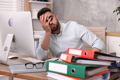 Overwhelmed man sitting at table with computer and stack of folders in office