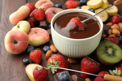 Photo of Fondue fork with strawberry in bowl of melted chocolate surrounded by other fruits on wooden table