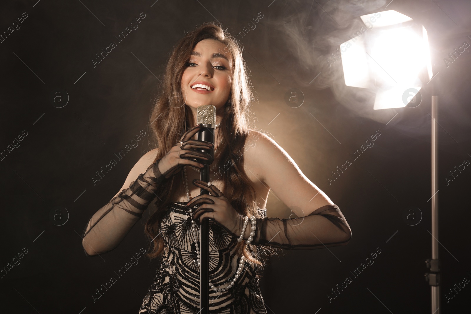 Photo of Beautiful young woman with microphone singing on dark background with smoke