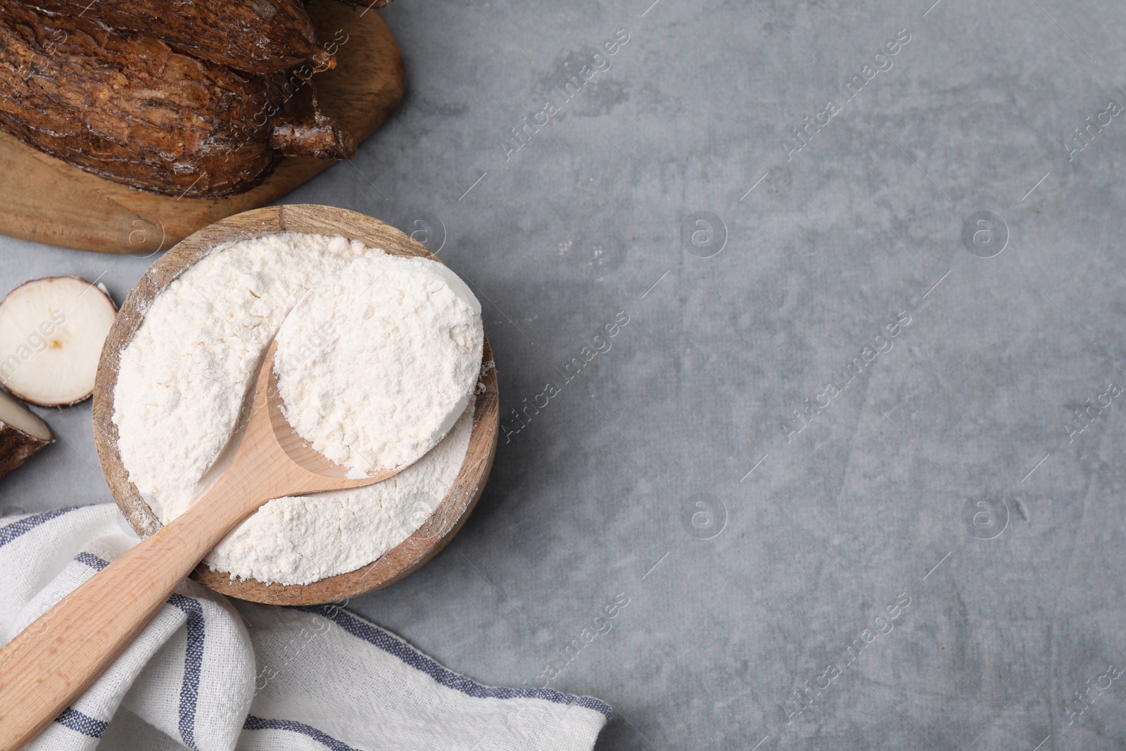 Photo of Wooden bowl, spoon with cassava flour and roots on grey table, flat lay. Space for text