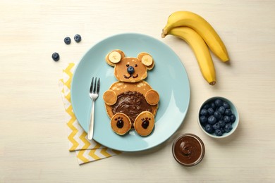 Photo of Creative serving for kids. Plate with cute bear made of pancakes, blueberries, bananas and chocolate paste on light wooden table, flat lay