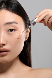 Photo of Beautiful young woman applying cosmetic serum onto her face on grey background, closeup