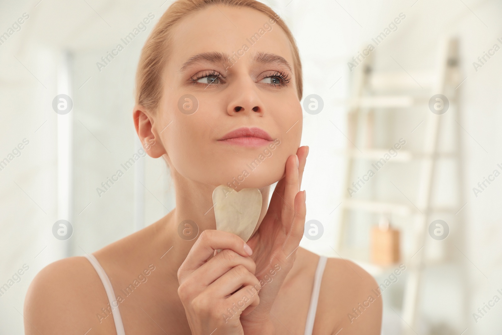 Photo of Beautiful young woman doing facial massage with gua sha tool in bathroom, closeup