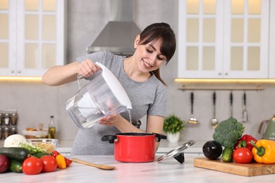 Photo of Happy young housewife pouring water in pot at white marble table in kitchen. Cooking process