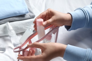 Photo of Cosmetic travel kit. Woman putting small bottle with personal care product into plastic bag on bed, closeup